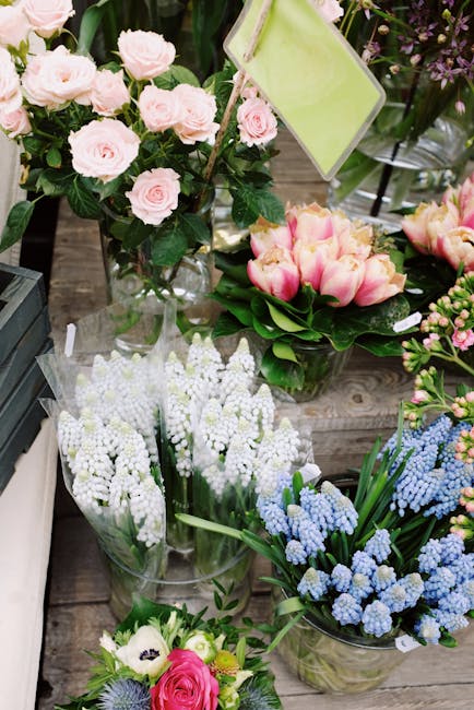 A display of fresh floral arrangements arranged on a rustic wooden surface, featuring a bouquet of pale pink roses with dark green foliage in a clear glass vase, a cluster of pink and yellow tulips wi